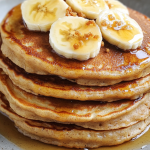 A stack of fluffy banana pancakes topped with fresh banana slices, brown sugar crumbles, and drizzled with maple syrup on a ceramic plate.