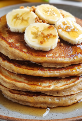 A stack of fluffy banana pancakes topped with fresh banana slices, brown sugar crumbles, and drizzled with maple syrup on a ceramic plate.